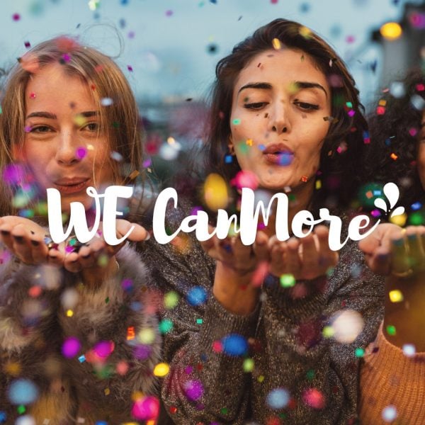 Two women blow a shower of coloured confetti straight at the camera, hair flying, with WE CanMore written in white script across the image. A women's gathering that does not take itself too seriously.